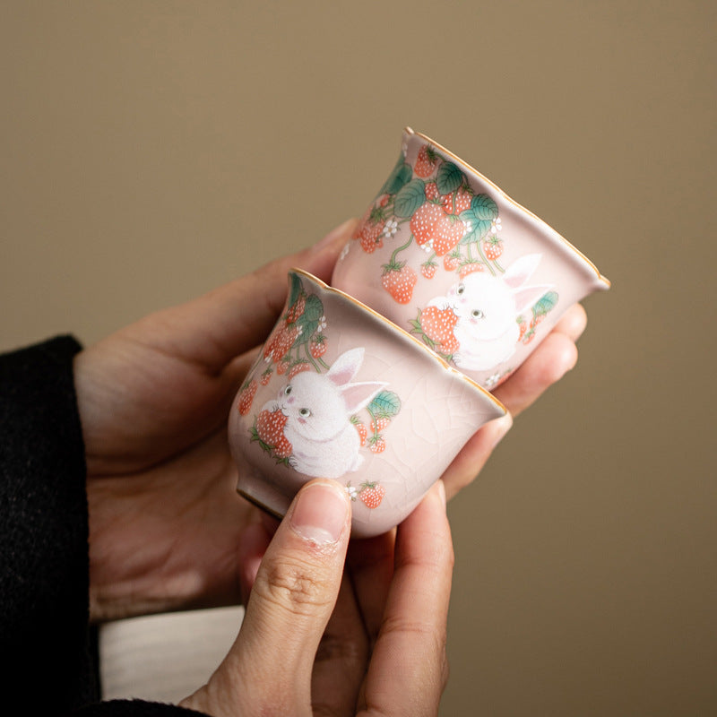 Two hands holding small pink ceramic cups with painted white rabbits and strawberries