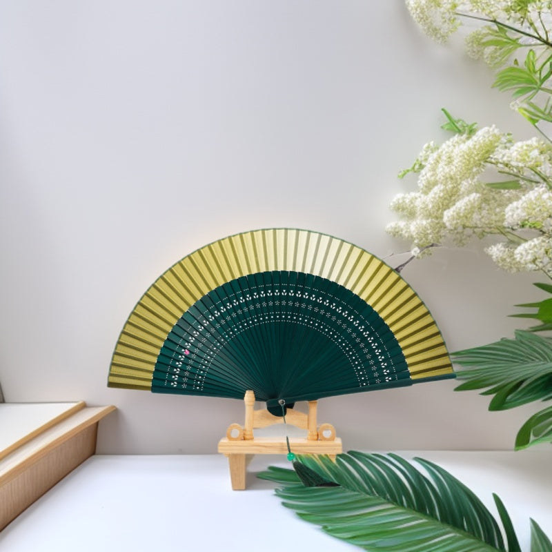 Green and yellow gradient handheld folding fan with hollow engravings displayed on a wooden stand