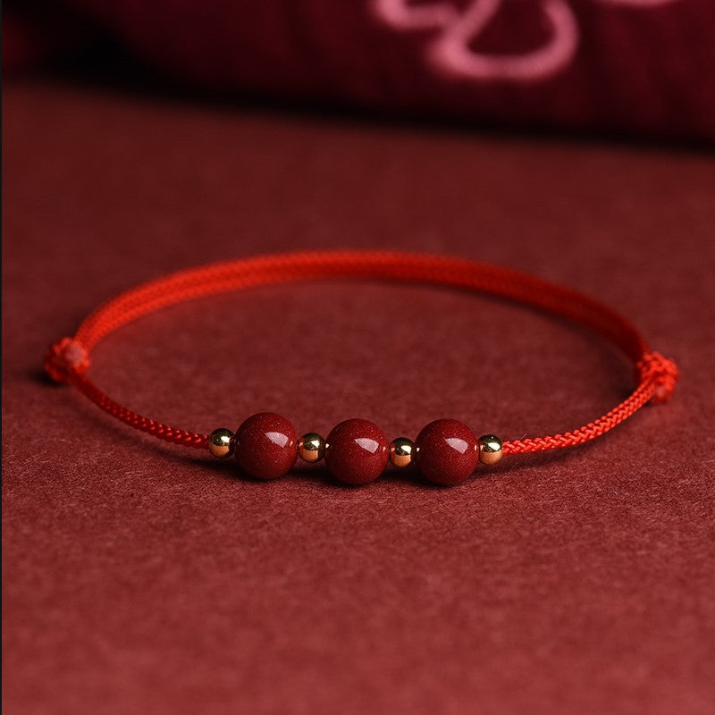 A close-up of a red braided cord bracelet featuring three round natural cinnabar beads separated by small gold-tone spacers.
