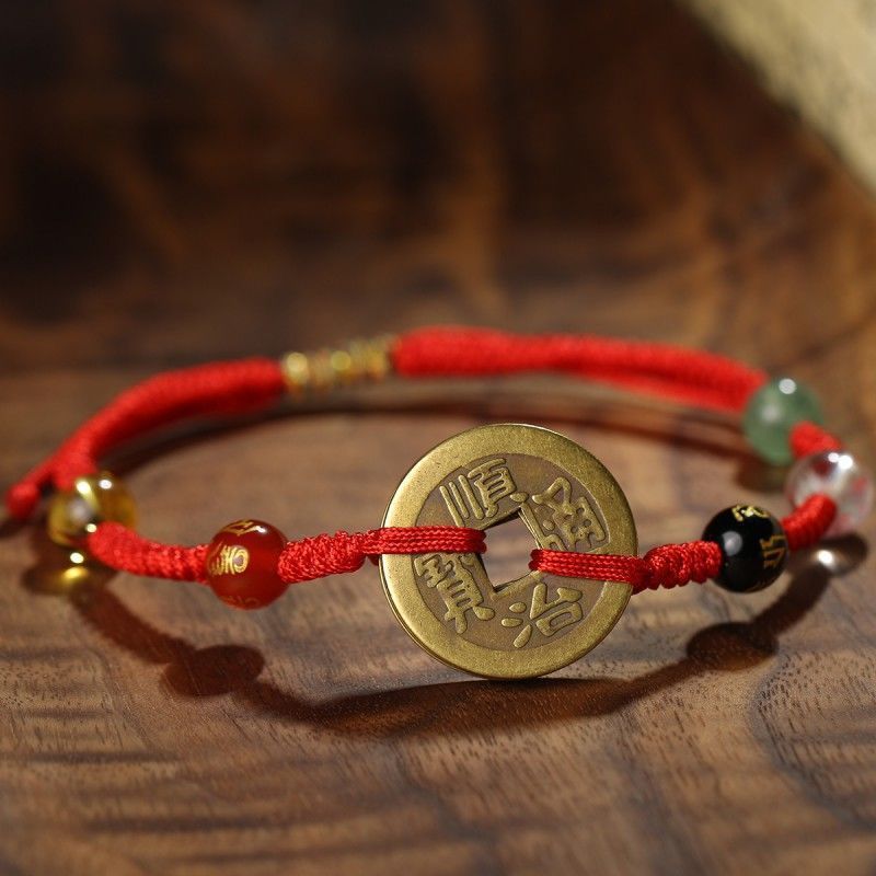 Close-up of a red string bracelet featuring a central copper coin charm and small accent beads on a wooden surface