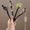 A hand holding four black wooden hairpins with different floral and leaf decorations and hanging beads