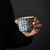 A hand holding a small blue and white porcelain teacup with Chinese calligraphy text.