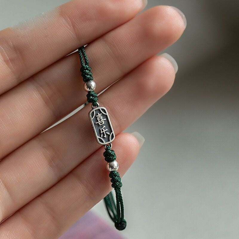 Hand holding a dark green braided bracelet with a rectangular metal charm engraved with Chinese characters.