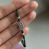 Hand holding a dark green braided bracelet with a rectangular metal charm engraved with Chinese characters.