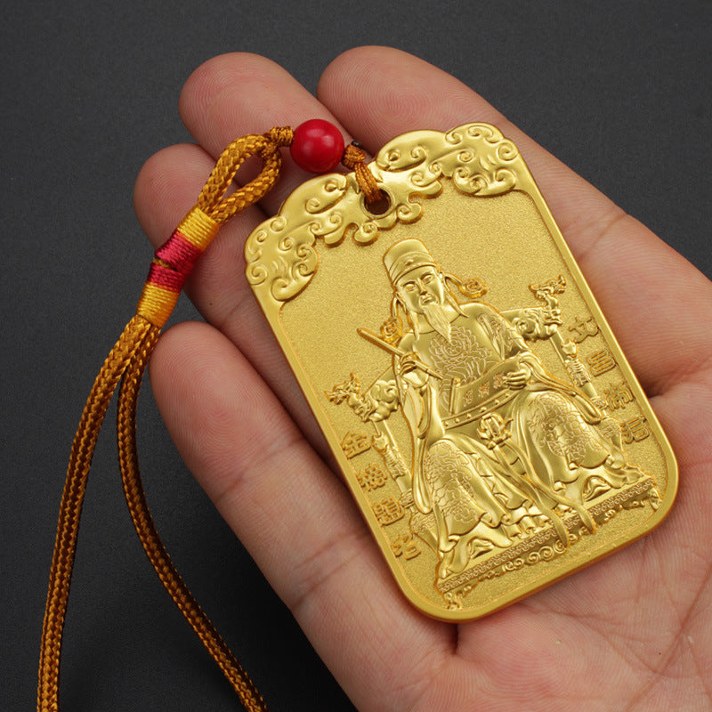 Hand holding a rectangular gold-tone metal pendant featuring a seated scholar figure