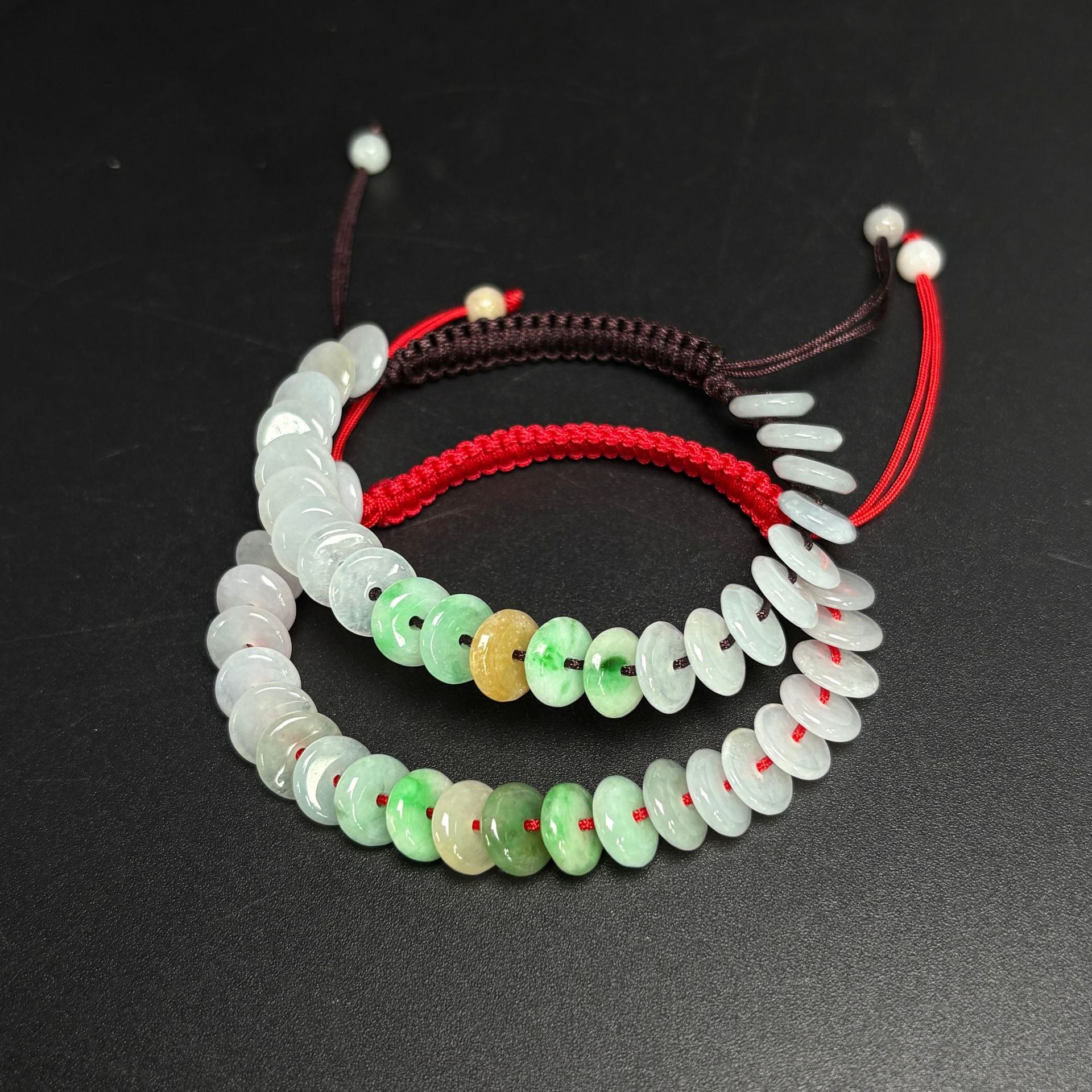 Two jade bracelets with round beads on red and brown woven cords lying on a black surface