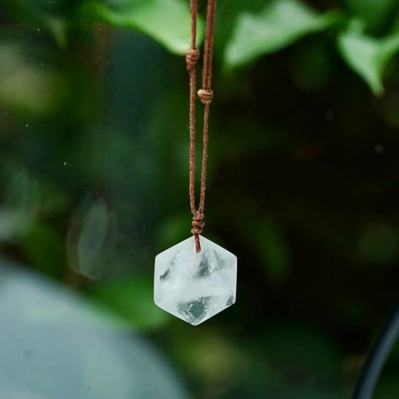 A clear white crystal hexagon pendant hanging on a brown cord against green leaves