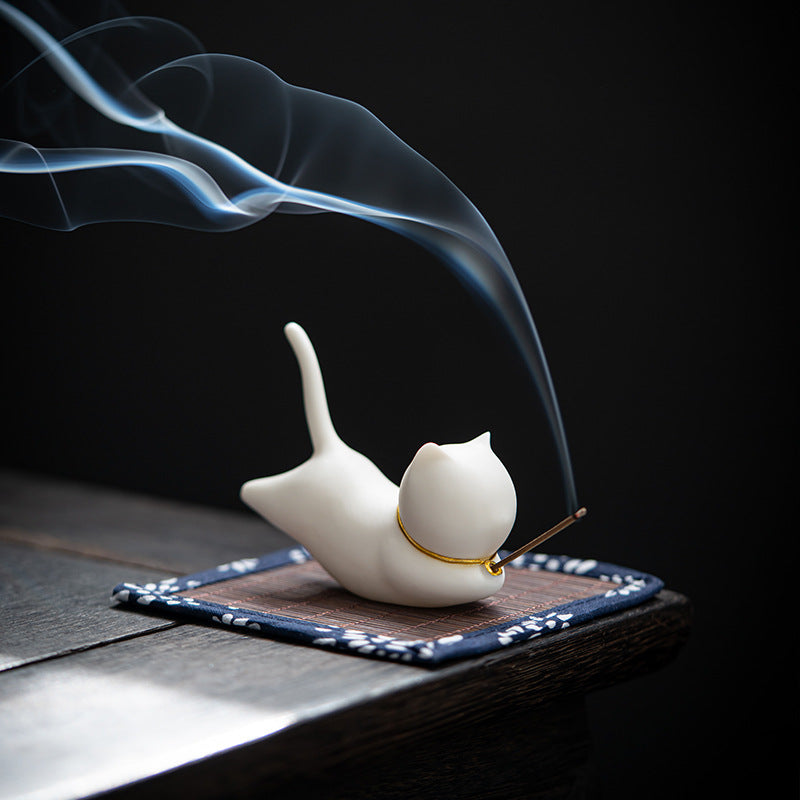 White ceramic cat incense burner holding an incense stick with smoke rising against a dark background