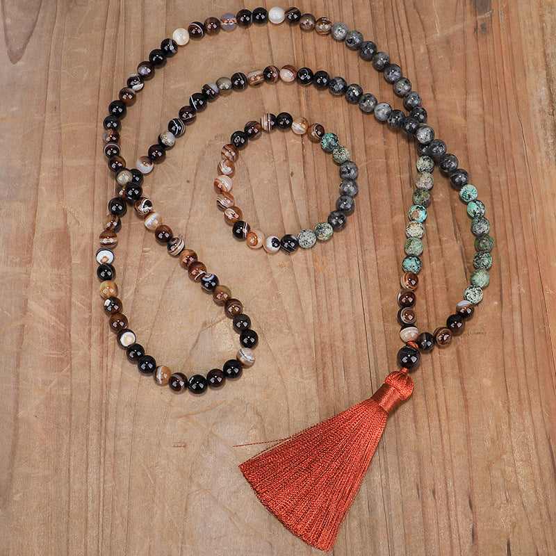 Long 108 bead mala necklace with mixed stones and an orange tassel next to a matching bracelet on wood