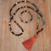 Long 108 bead mala necklace with mixed stones and an orange tassel next to a matching bracelet on wood