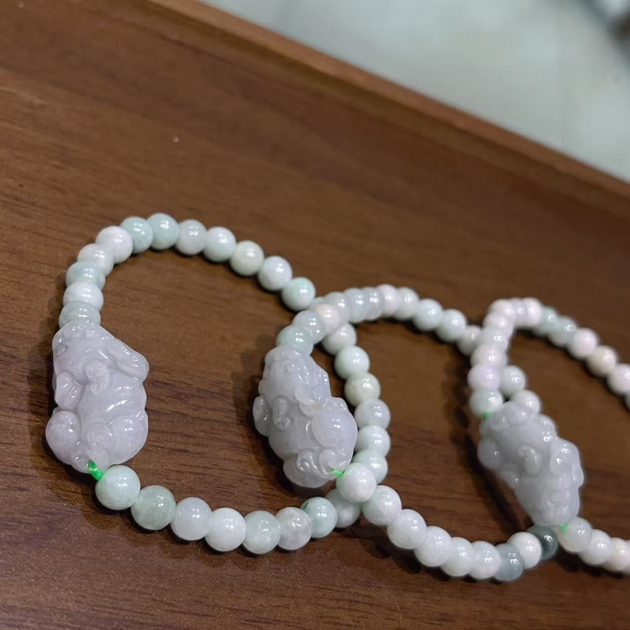 A close-up of three pale green jade bracelets with carved Pixiu charms resting on a wooden surface.