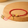Close up of a red woven bracelet with a gold flower charm and green jade beads on a wooden surface