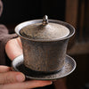 Hand holding a rustic brown ceramic gaiwan teacup with lid and saucer