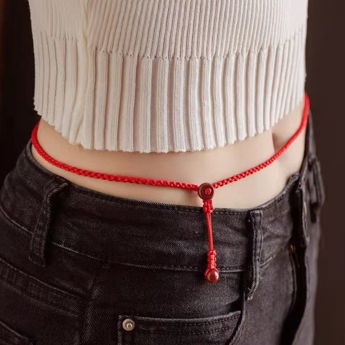 Model wearing a red woven cord waist chain with a hanging cinnabar bead over dark jeans