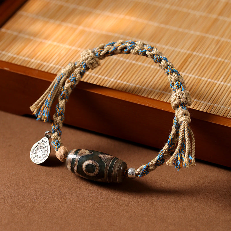 Close up of a hand-woven bracelet with a brown patterned Dzi bead and silver charm on a bamboo mat