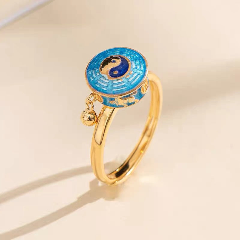 Close-up of a gold-tone copper ring with a blue enamel Yin Yang top and a small dangling bell.