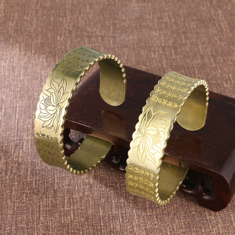 Two brass open cuff bracelets with lotus flower and Heart Sutra engravings resting on a wooden stand